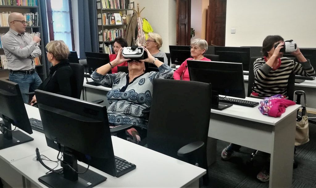 Grupa seniorów siedzi przy stolikach z komputerami, niektórzy nich mają na oczach gogle do oglądania wirtualnej rzeczywistości.