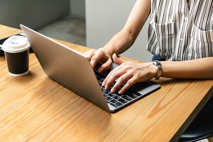 Fragment blatu stolika, na którym stoi laptop i kubek. Przy stoliku kobieta pracuje na laptopie. Widać tylko jej ręce i fragment tułowia