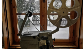 Na tle okna z zimowym pejzażem na zewnątrz, projektor filmowy w kolorze szarym z pustą rolką na taśmę filmową
